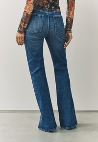 Modré džínsové nohavice s rozšíreným strihom, stredne prané, s päťvreckovým dizajnom a miernym elastanom. Vyznačujú sa viditeľným prešitím a hladkou textúrou.