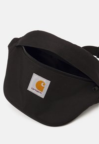 Czarna torba na pas z materiału o zaokrąglonym kształcie, z zamkiem błyskawicznym i białą łatą z pomarańczowym logo. Gładka faktura, minimalistyczny design.