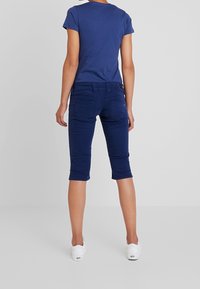 Osoba v modrém přiléhavém tričku a tmavě modrých kalhotách ke kolenům, stojící bosá proti jednobarevnému světlému šedému pozadí.