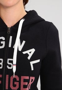 Svart zip-up hoodie med vita dragsko. Framsidan har stor vit och vinröd text. Tillverkad av mjuk tyg med avslappnad passform.