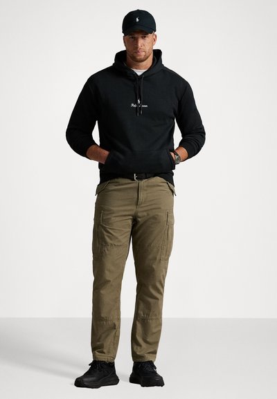 Mężczyzna w czarnej bluzie Ralph Lauren, czapce w kolorze czarnym, oliwkowych spodniach cargo i czarnych trampkach, stojący z rękami w kieszeniach na jednolitym tle.