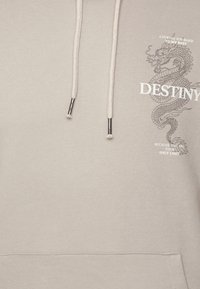 Sudadera de color gris claro con un gráfico de dragón y el texto "DESTINO." Cuerdas con puntas de metal y una tela suave y texturizada.