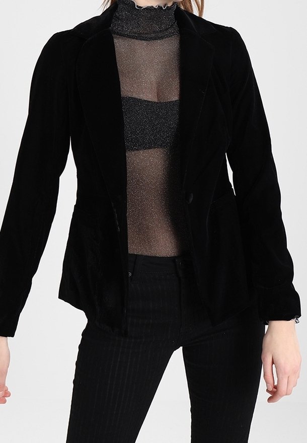 Personne portant un blazer en velours noir par-dessus un top noir transparent pailleté et un pantalon noir, debout devant un fond clair uni.
