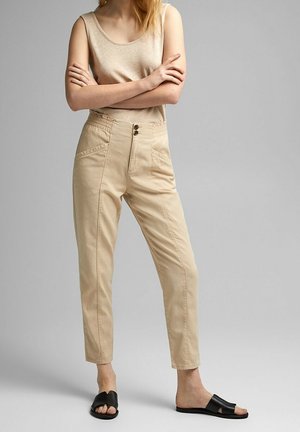 Kobieta stojąca z założonymi skrzyżowanymi ramionami, ubrana w beżową bluzkę bez rękawów, beżowe spodnie z wysokim stanem oraz czarne sandały wsuwane na szarym tle.