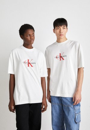 Camisetas de algodón blancas con el gráfico rojo "Calvin Klein Jeans", de manga corta, corte relajado, combinadas con pantalones casuales negros y azul claro.