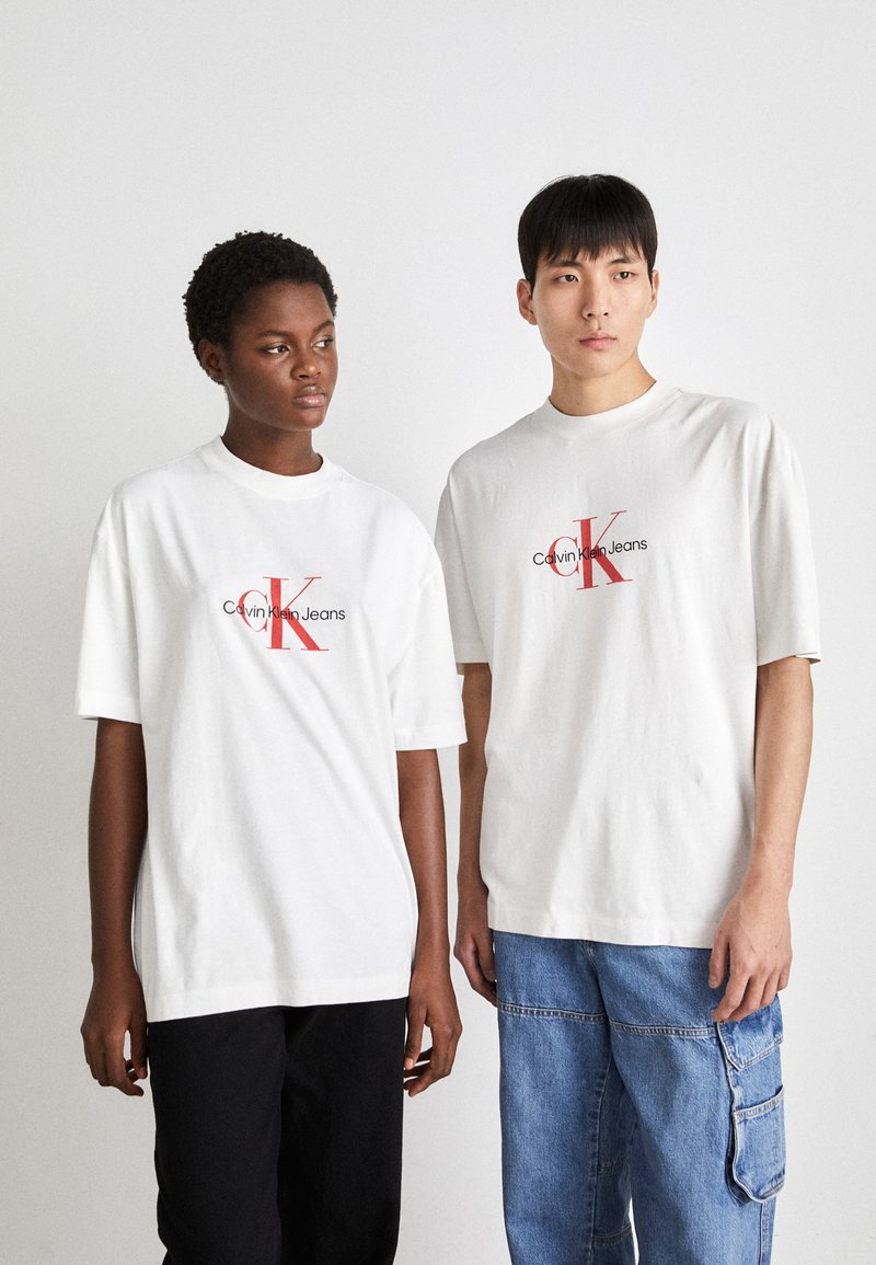 Bílé bavlněné trička s červenou grafikou "Calvin Klein Jeans", krátké rukávy, volný střih, v kombinaci s černými a světle modrými volnými kalhotami.