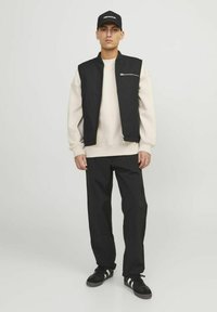 Sort ærmeløs vest over en beige sweatshirt, kombineret med sorte bukser og sorte sneakers med hvide striber. En kasket fuldender looket.