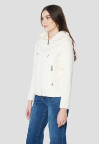 Fehér, steppelt kapucnis dzseki cipzáros elülső résszel, két oldalzsebbel és kontrasztos, sima ujjakkal. Kék farmernadrág fölött viselve.