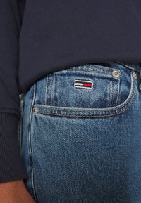 Blå denimjeans med en blekt textur, med en liten logotypapplikation i röd, vit och svart på bakfickan.
