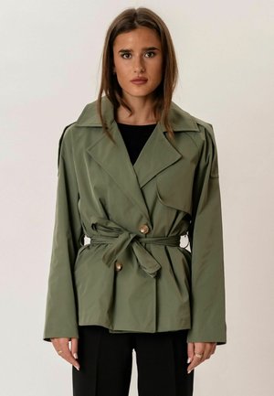 Fiatal nő olívazöld öves dzsekit visel fekete felső és nadrág fölött, sima világos háttér előtt áll.