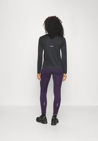 Femme se tenant de dos, portant un haut noir à manches longues, des leggings violets avec des bandes réfléchissantes, des chaussettes noires et des chaussures de course noires.