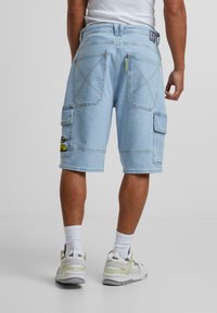 Lyse blå denim cargo shorts med flere lommer, synlige sømdetaljer og et merkeetikett på livet. Brukt med hvite sokker og sko.