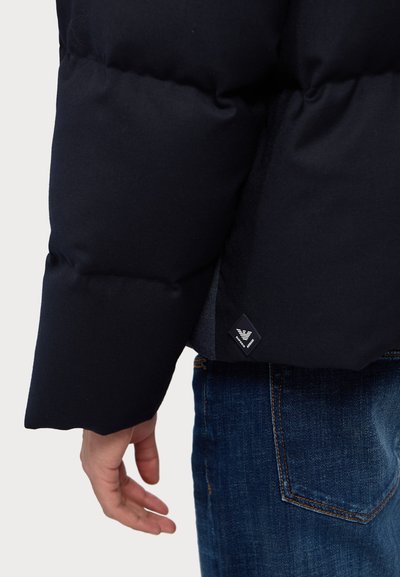 Navy padded jacket with a textured finish, featuring a logo tag on the bottom and a relaxed fit, paired with blue denim jeans.