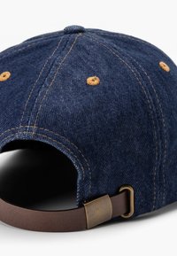 Jeanskeps i marinblå färg med brun läderrem och mässingsspänne. Har gula broderidetaljer runt ventilationshålen.