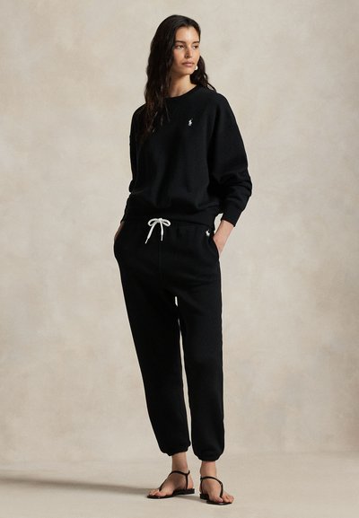 Γυναίκα που στέκεται με τα χέρια στις τσέπες, φορώντας μαύρο πουλόβερ με μακριά μανίκια και ταιριαστό παντελόνι jogger με λευκό κορδόνι, καθώς και μαύρα σανδάλια.