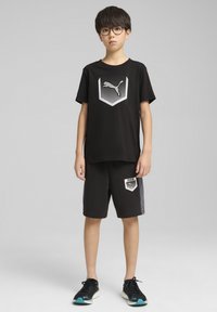 Camiseta negra con logo plateado de Puma, combinada con pantalones cortos negros con detalles grises y logo. Zapatillas deportivas negras con detalles en teal.