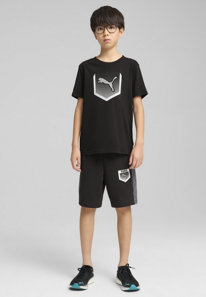 Camiseta negra con logo plateado de Puma, combinada con pantalones cortos negros con detalles grises y logo. Zapatillas deportivas negras con detalles en teal.