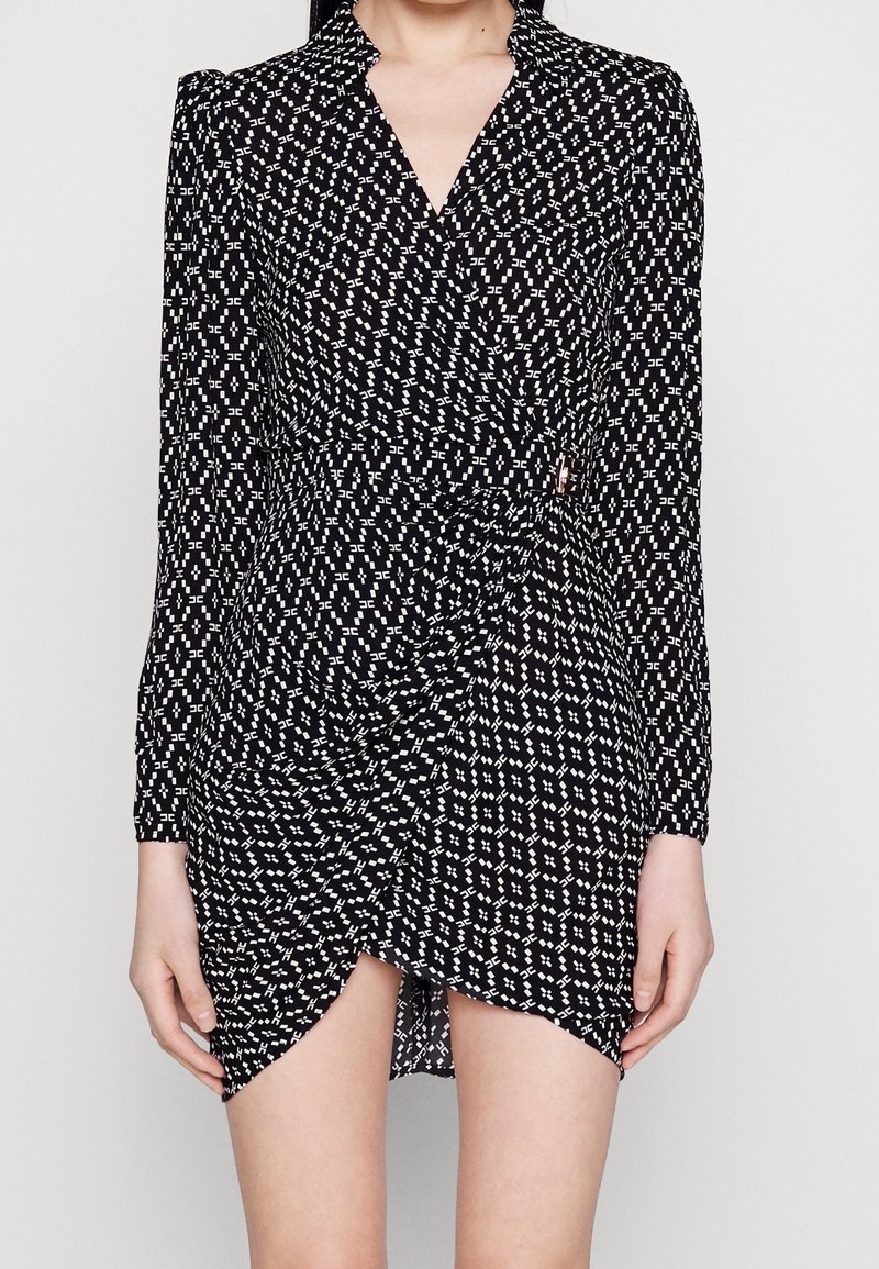 Femme portant une robe portefeuille noire à manches longues avec un motif géométrique blanc et un ourlet asymétrique dévoilant le haut des jambes.