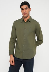 Μακρυμάνικο πουκάμισο σε ελιά πράσινο με γιακά με κουμπιά, κουμπιά μπροστά και διακριτικό λογότυπο στο στήθος· απαλό ύφασμα, στενή γραμμή.