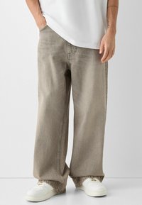 Jeans de pierna ancha en gris claro, fabricados en denim, con un acabado desgastado, costuras visibles y bolsillos, combinados con zapatillas blancas.