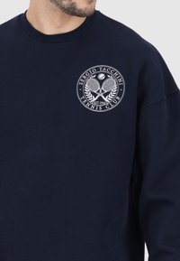 Felpa blu marina con scollatura rotonda. Presenta un logo ricamato bianco con racchette da tennis e foglie di alloro. Tessuto misto cotone.