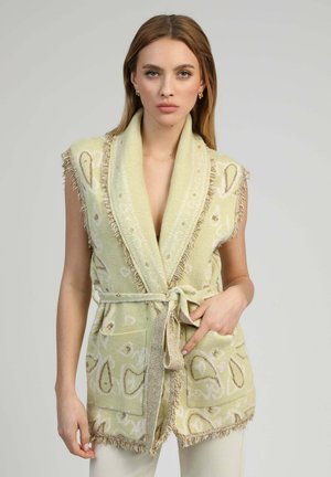Femme portant un gilet en tricot sans manches beige à motif cachemire avec ceinture, debout devant un fond gris clair uni.