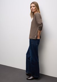 Donna in piedi di profilo con le mani nelle tasche, che indossa un maglione marrone con dettagli di bottoni e jeans a gamba larga blu scuro con bottine.