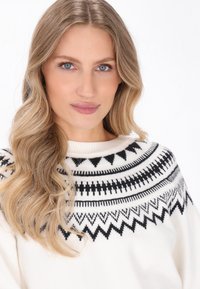 Maglione bianco con motivo a maglia geometrica nera sul colletto. Tessuto morbido con un ampio colletto a costine. Lunga chioma fluente visibile.