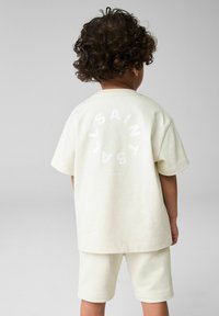 Krémfarvet bomulds T-shirt med korte ærmer og ryggrafik med teksten "SAINT LONDON" i hvid cirkulær skrift. Bærer matching shorts.