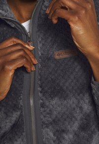 Fleecejacka i mörkgrått med en texturerad, plyschigare yta, med dragkedja fram och en brun läderlogotyp på bröstet.