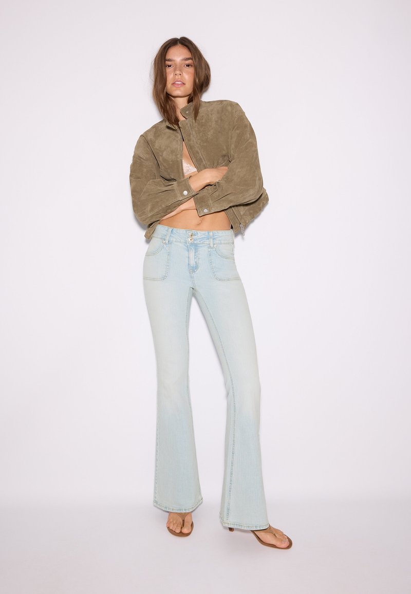 Femme portant une veste olive courte, un jean flare taille haute bleu clair et des sandales à talons marron sur un fond blanc uni.