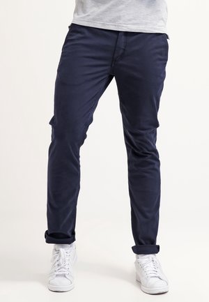 Mężczyzna w granatowych, dopasowanych spodniach i białych trampkach stojący na jasnym tle, z lekko rozstawionymi nogami i podwiniętymi nogawkami.