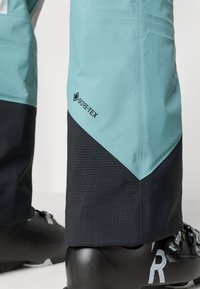 Wodoszczelne spodnie narciarskie w kolorze turkusowym i czarnym, z metką Gore-Tex, kanciastym blokowaniem kolorów oraz gładką teksturą.