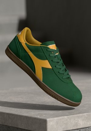 Green and yellow athletic shoe made of suede, featuring a textured sole, rounded toe, and lace-up design with a prominent side logo.