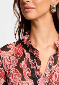 Rood en zwart gemêleerd chiffon blouse met een gerimpelde kraag en knoopsluiting. Grote zilveren oorring zichtbaar bij het model.