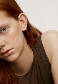 Pendiente de aro dorado con un delicado detalle de cadena, que se lleva en la oreja izquierda. El sujeto tiene el cabello largo y ondulado de color rojo y viste una camiseta marrón.