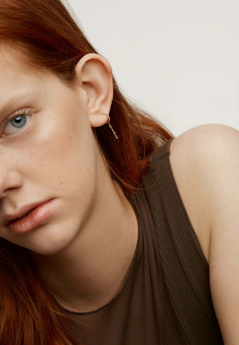 Pendiente de aro dorado con un delicado detalle de cadena, que se lleva en la oreja izquierda. El sujeto tiene el cabello largo y ondulado de color rojo y viste una camiseta marrón.