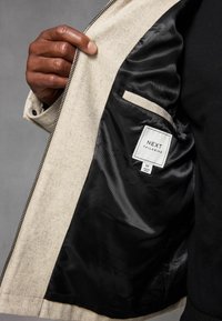 Beżowa kurtka o gładkim, teksturowanym wykończeniu i czarnym podszewką. Posiada wewnętrzną kieszeń oraz metkę z napisem "NEXT TAILORING."