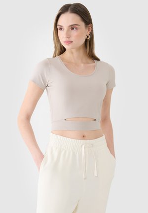 Młoda kobieta ubrana w beżowy krótki crop top z wycięciem z przodu, zestawiony z białymi spodniami z wysokim stanem i sznurkiem, stoi z rękami w kieszeniach.
