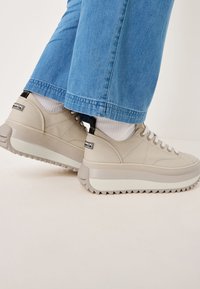 Sneakers in tela beige con suola grigia spessa e rigata, lacci bianchi e un tappo sul tallone nero. Superficie texture con dettagli delle cuciture.