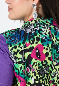 Bunte Jacke mit einem lebhaften Muster aus Herzen, Tierdrucken und auffälligem Text auf grünem Hintergrund; lila Ärmel. Silberne Statement-Ohrringe.