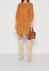 Abito in corduroy color ruggine con chiusura a bottoni, maniche a sbuffo e vita elastica. Abbinato a stivali beige al ginocchio e una borsa marrone.