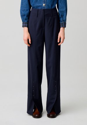 Persona che indossa pantaloni larghi blu navy con spacchi e bottoni frontali, abbinati a una camicia di denim e scarpe marroni.