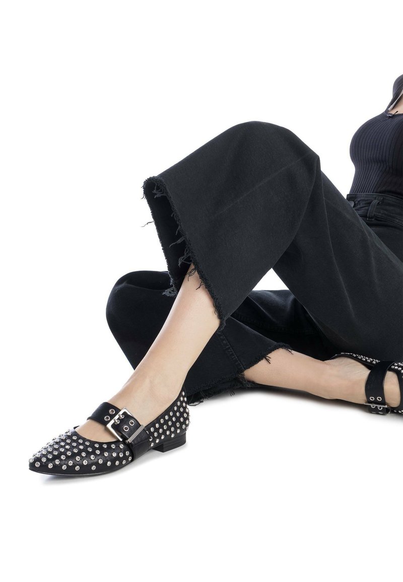 Bailarinas negras con tachuelas y punta afilada, con tira de hebilla y superficie texturizada, combinadas con jeans negros de pierna ancha con dobladillos deshilachados.