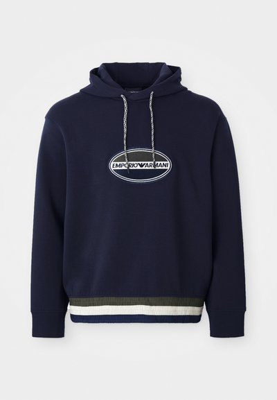 Sudadera azul marino con logo bordado en un parche ovalado. Cuenta con capucha ajustable y un dobladillo acanalado texturizado con rayas contrastantes.