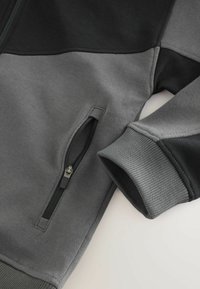 Veste zippée grise et noire au design épuré, avec une texture de tissu lisse et des poignets côtelés. Poche latérale visible.