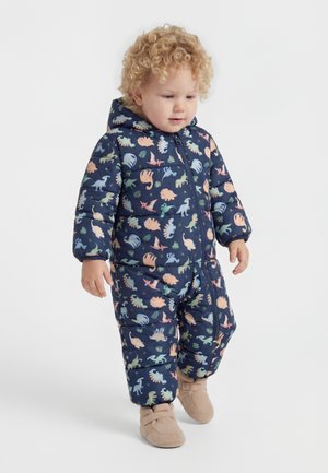 Göndör hajú kisgyerek sötétkék, dinoszaurusz mintás kapucnis hóruhába és bézs színű puha papucsba öltözve áll fehér háttér előtt.