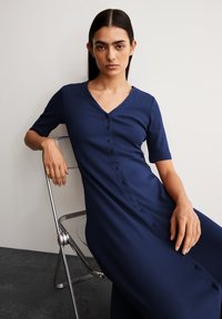 Robe midi côtelée bleu marine avec des manches courtes et un décolleté en V, dotée d'un devant à boutons et d'une texture lisse. Modèle assis sur une chaise argentée.