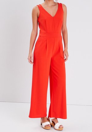 Frau trägt einen ärmellosen roten Jumpsuit mit weitem Bein und V-Ausschnitt sowie weiße Plateausandalen, steht vor hellgrauem Hintergrund.