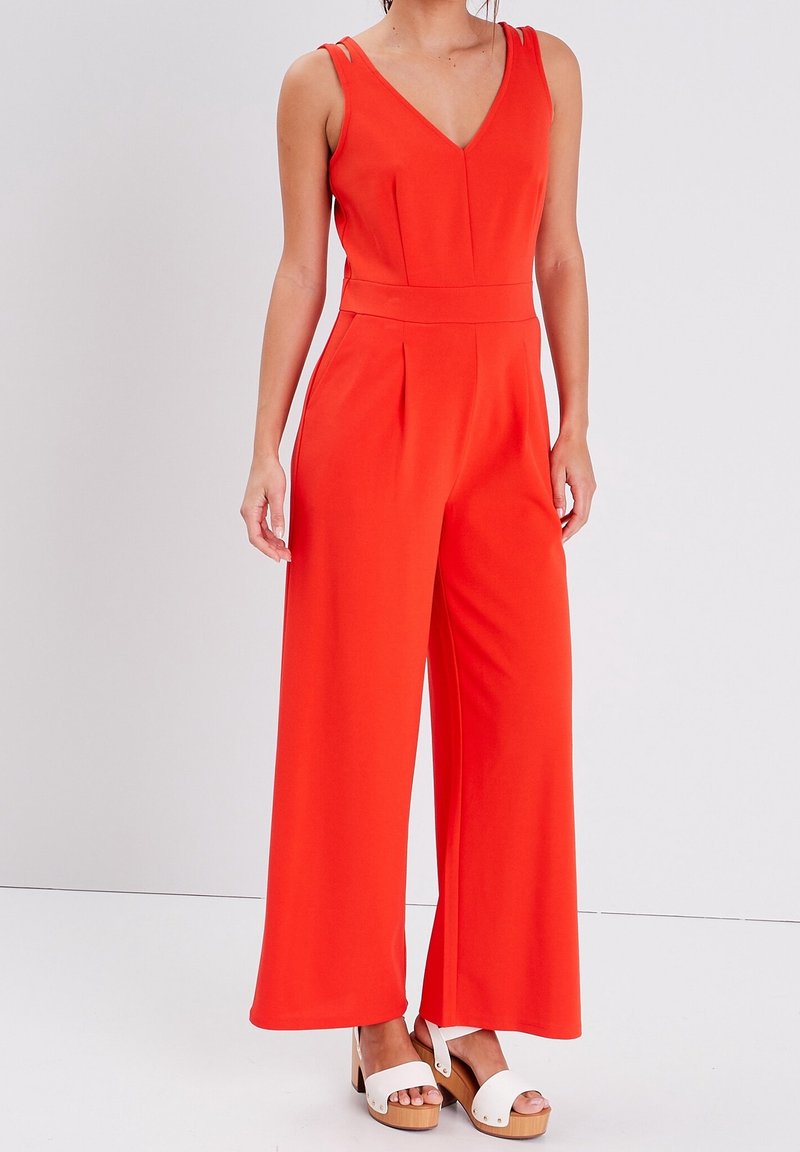 Femme portant une combinaison rouge sans manches à jambes larges avec un col en V et des sandales blanches à plateforme, debout devant un fond gris clair.